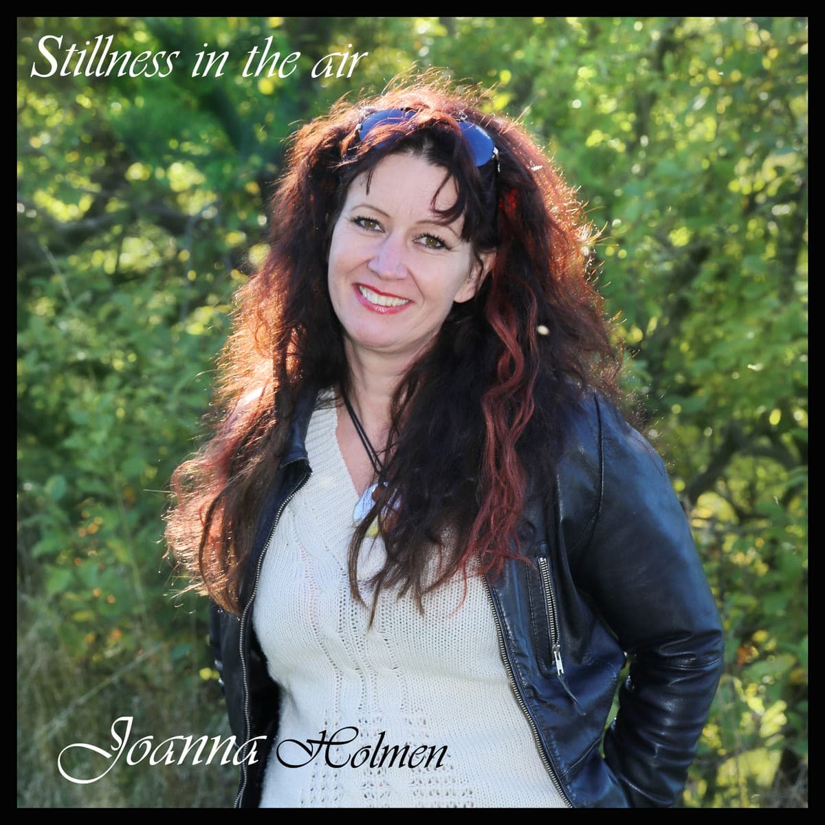 Joanna Holmen smiler i sort læderjakke og lys strik foran solbeskinnede træer. Cover til singlen Stillness in the air